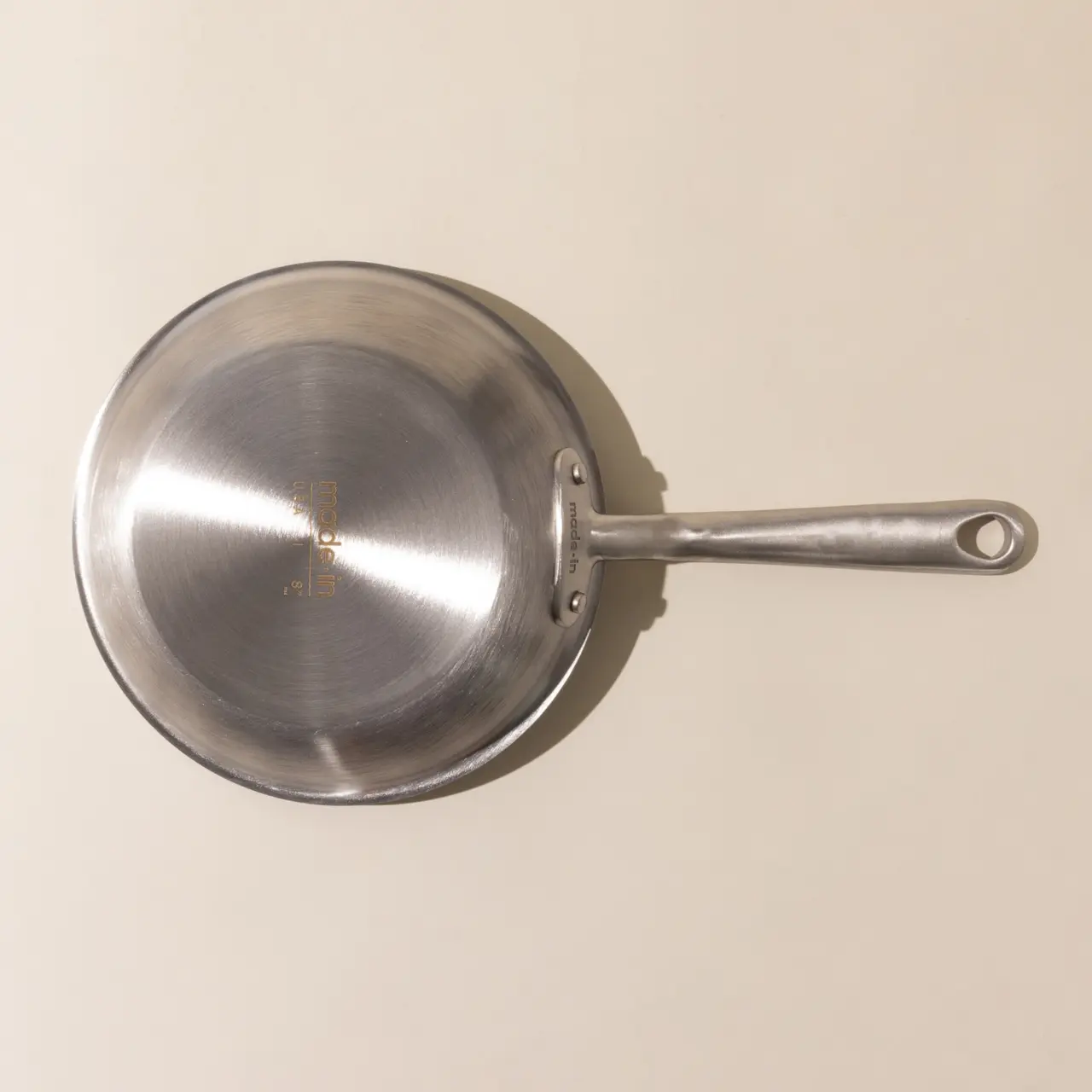 A stainless steel frying pan with a long handle on a beige surface viewed from above.