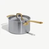A stainless steel pot with a lid and golden handles sits on a light background.