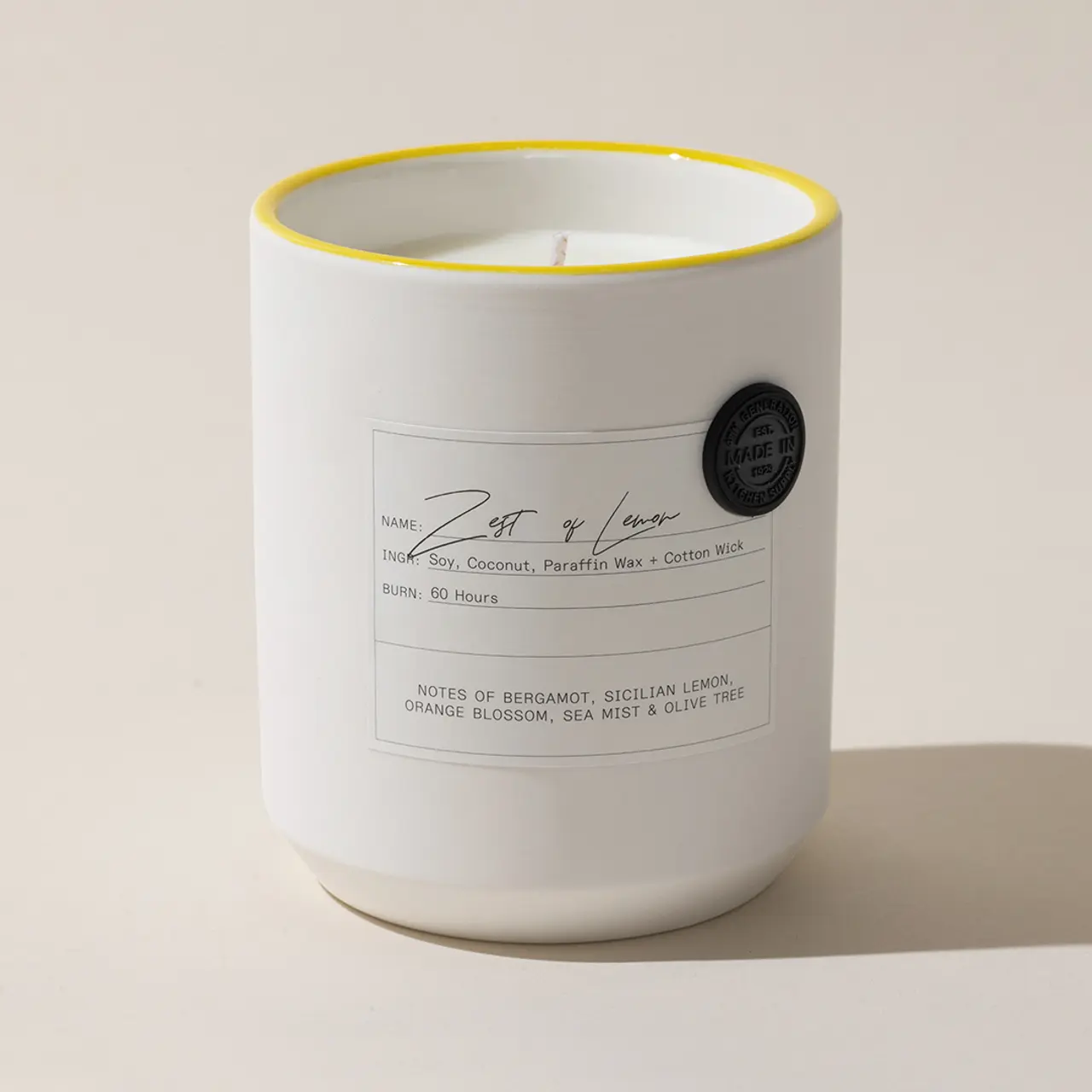 A lit scented candle with a yellow rim and label detailing its fragrance notes and ingredients is displayed against a plain background.