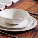 A stack of white ceramic bowls and plates is arranged neatly on a wooden table, accompanied by stainless steel utensils.