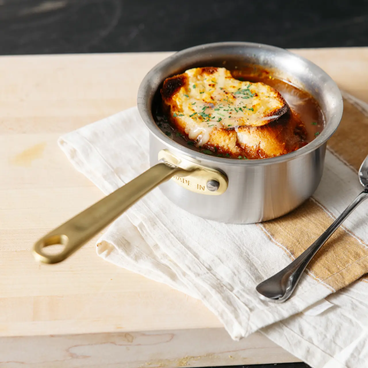 Made In Stainless Clad Butter Warmer in Antique Brass holds a bubbling, cheesy dish on a cutting board.