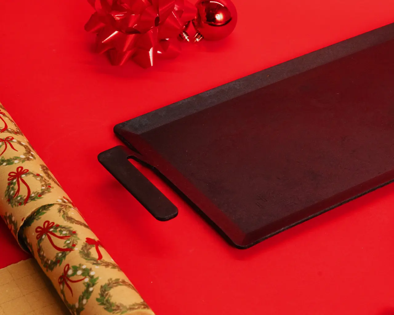 A black baking tray is surrounded by festive holiday decorations, including a red ornament and gift wrap featuring Christmas motifs on a red surface.