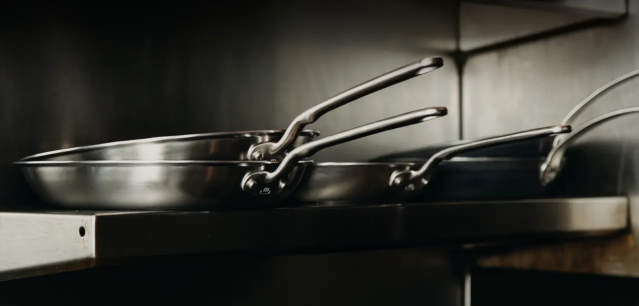 Three stainless steel frying pans with metal handles are stacked on a stove top against a dark background.