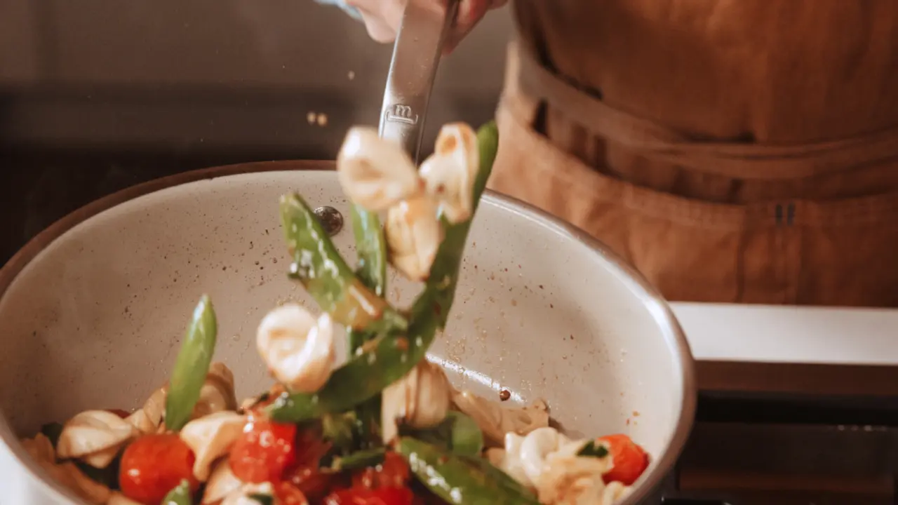 A hand is stirring a colorful mixture of tortellini, cherry tomatoes, and green vegetables in a large pot.