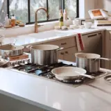 A modern kitchen countertop features various stainless steel pots and pans on a stove, with a bright window showcasing greenery outside.