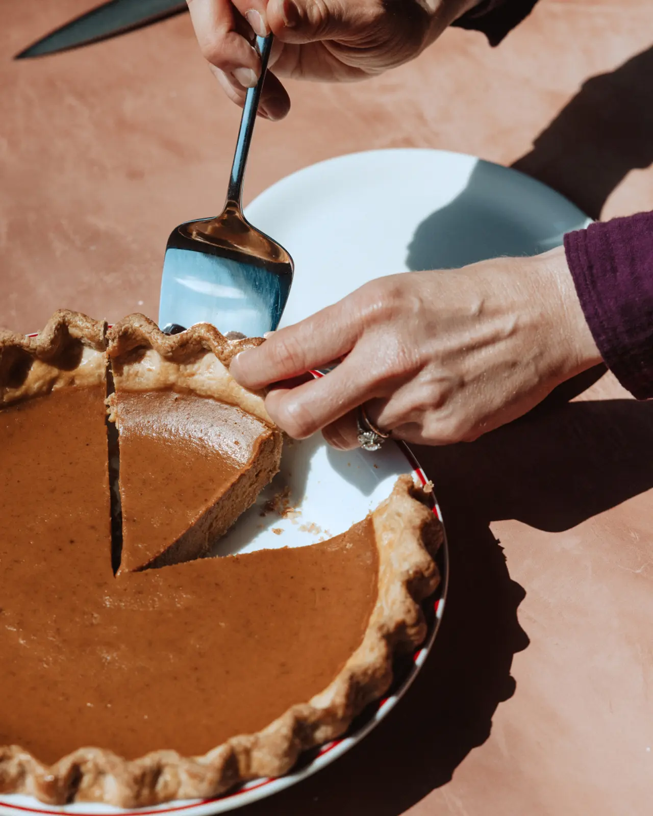 A hand is using a serving spoon to lift a slice from a pumpkin pie on a rustic table.