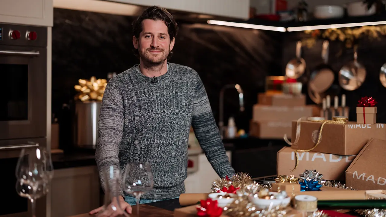 A man stands smiling at a table adorned with holiday decorations and gift-wrapping materials in a festive kitchen setting.