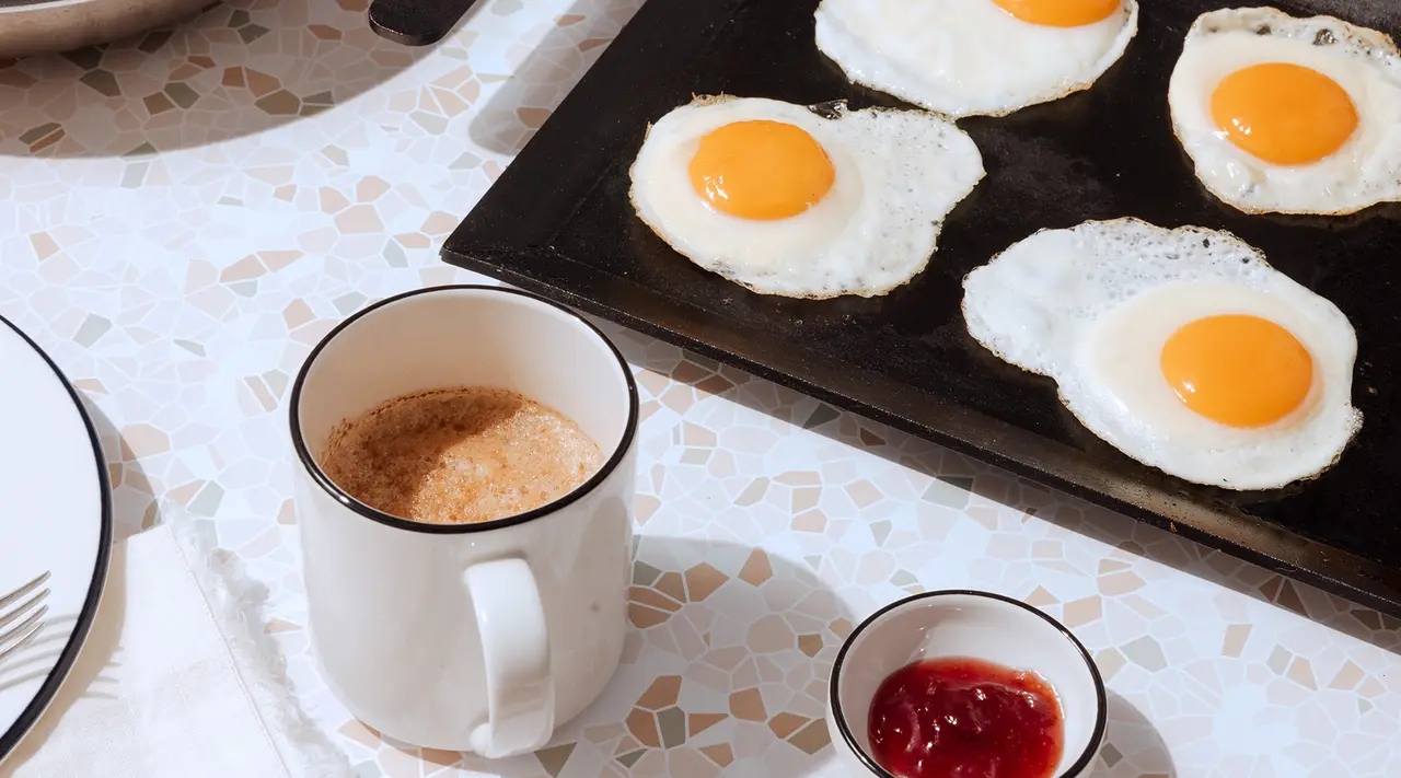 Fried eggs with bright yellow yolks are cooking on a black skillet, accompanied by a cup of coffee and a small bowl of ketchup on a textured table surface.