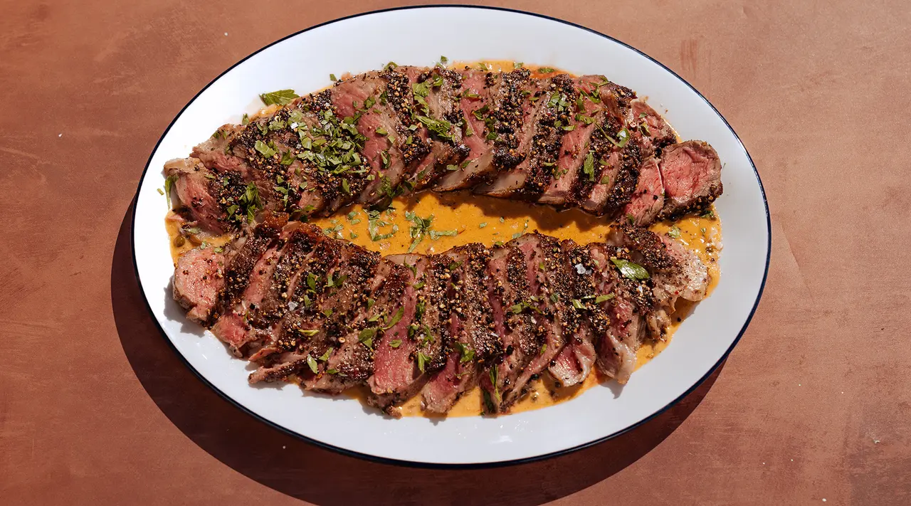 Sliced, medium-rare steak seasoned with herbs is served on an oval plate with a sauce.