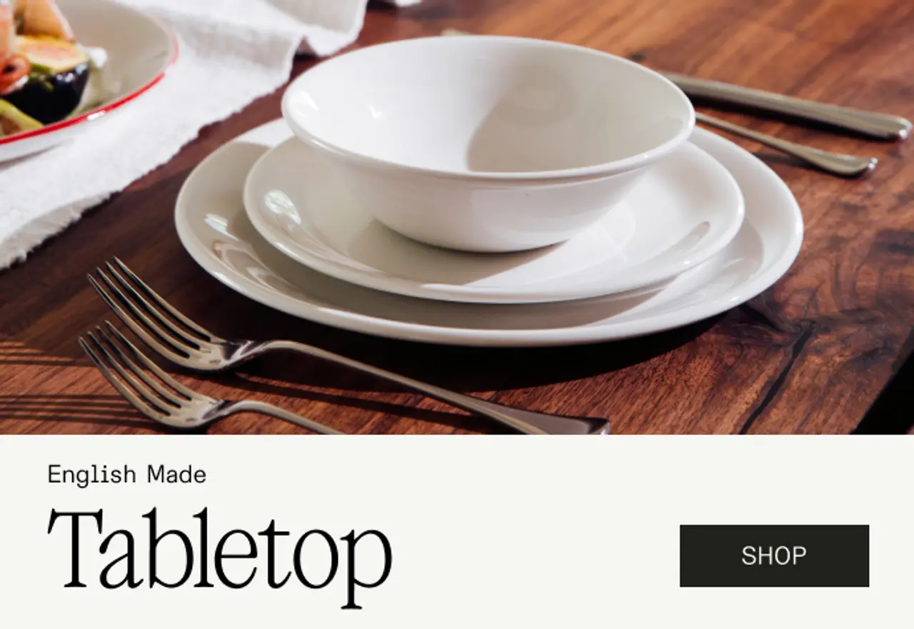Three elegantly stacked white ceramic plates and a bowl are arranged on a wooden table, accompanied by silverware, with a banner promoting an English-made tabletop collection.