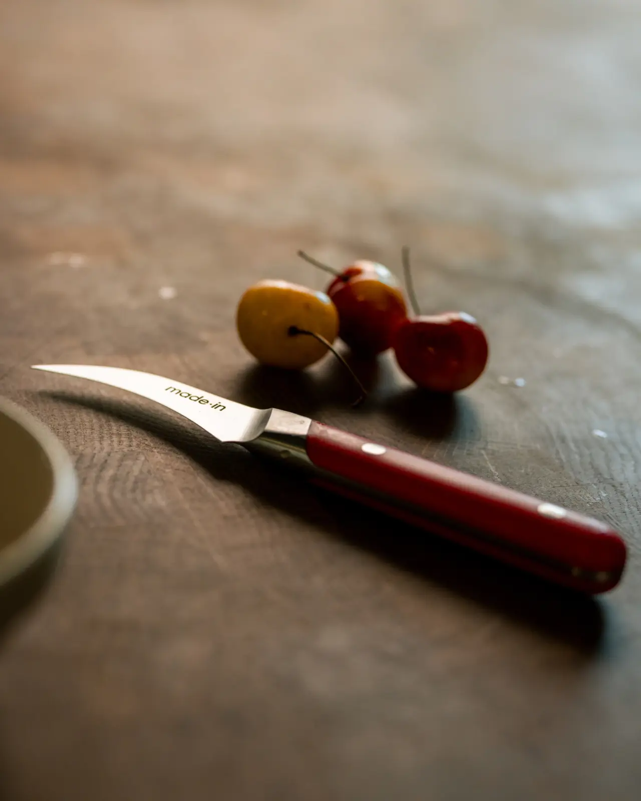 A small, curved knife with a red handle sits on a wooden surface next to three colorful fruit.