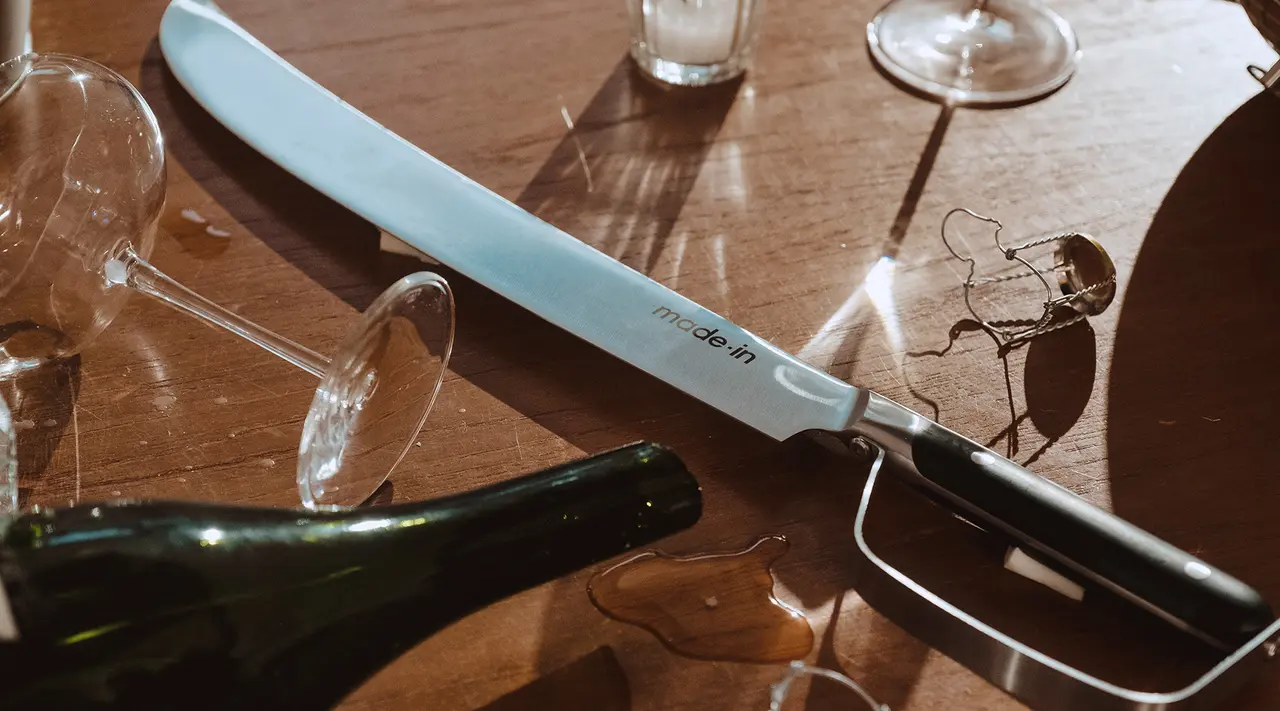 A long, flat knife with the words "made in" on the blade rests on a table surrounded by spilled wine and broken glassware.