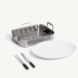 A stainless steel roasting pan with a rack is displayed alongside a large white serving plate and two kitchen knives.