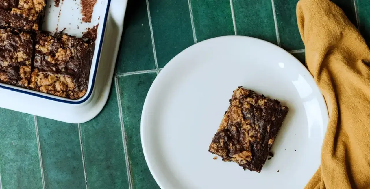 A single square of brownie is placed on a white plate next to a baking dish with more brownies and a yellow fabric napkin on a green tiled surface.