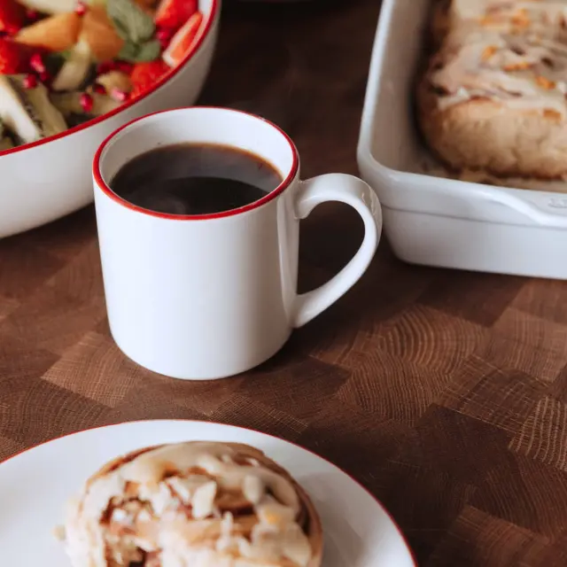A cozy breakfast scene featuring a white mug of coffee, a cinnamon roll, a bowl of fresh fruit, and a dish of baked goods on a wooden table.