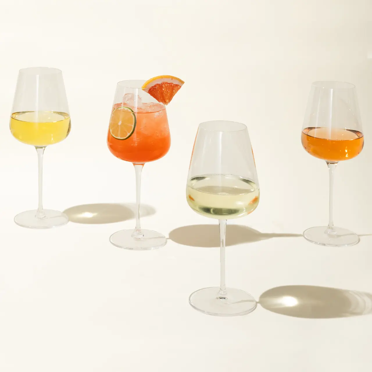 Four wine glasses with varying shades of beverages are lined up casting shadows on a light surface.