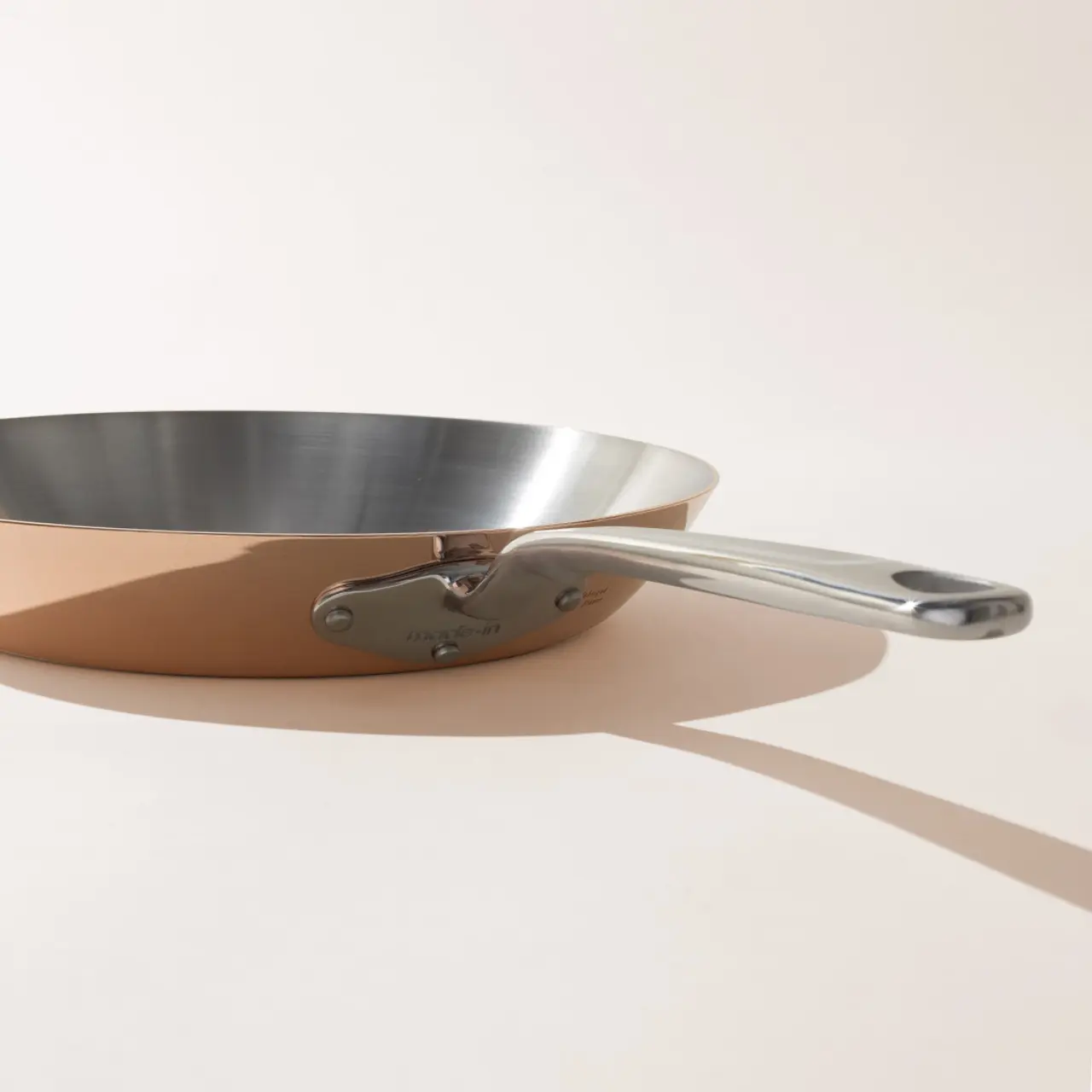 A stainless steel frying pan with a long handle casts a shadow on a light-colored surface.