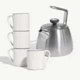 A stack of three white mugs with blue rims stands beside a sleek silver teapot.