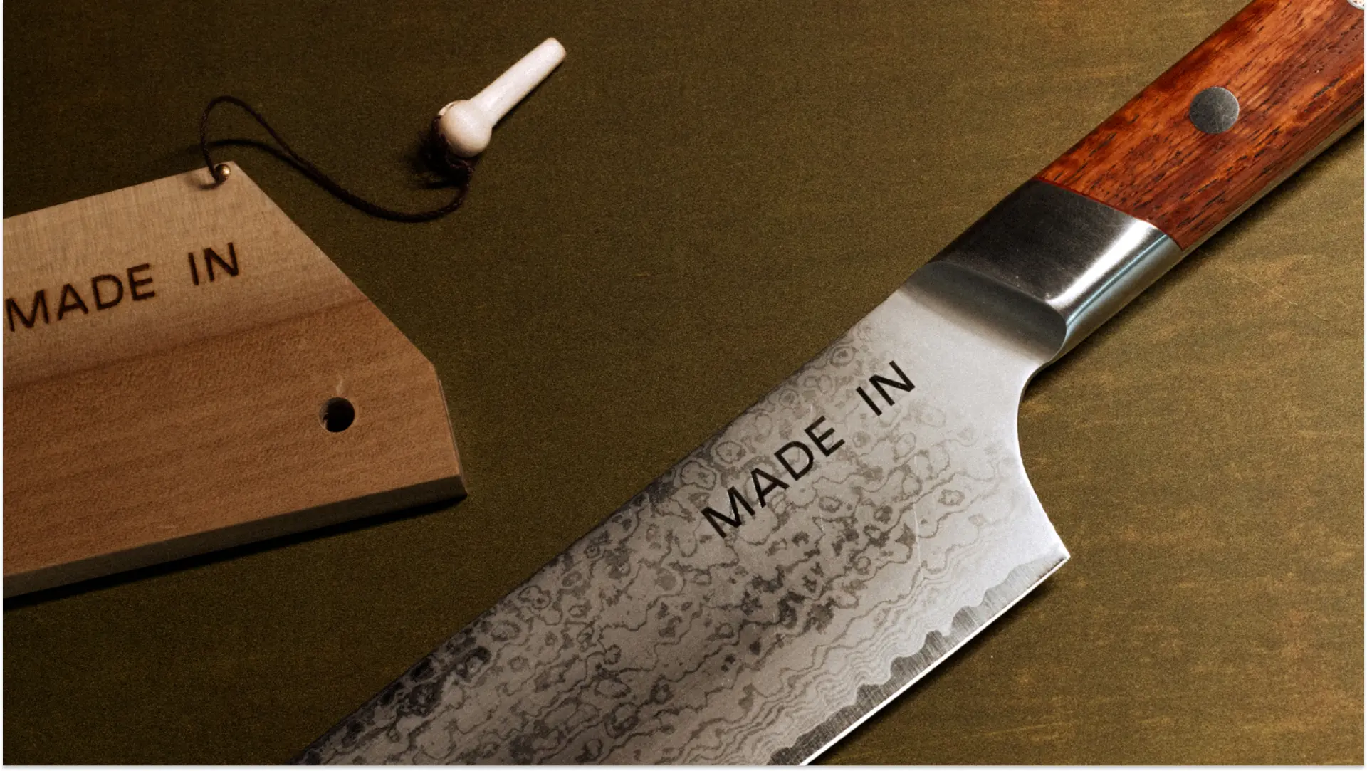 A close-up of a kitchen knife with a wooden handle and a patterned steel blade, next to a hanging tag marking it as "MADE IN."