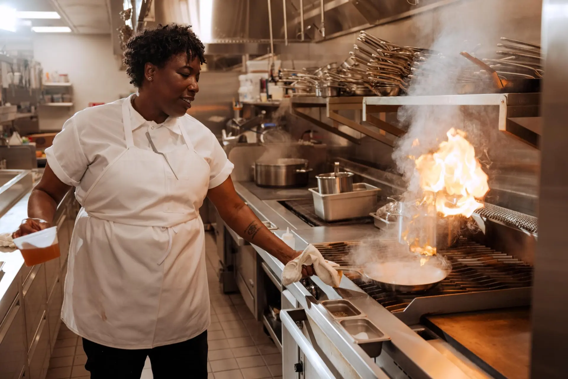 A chef skillfully manages a small flame erupting from a cooking pot in a busy kitchen.