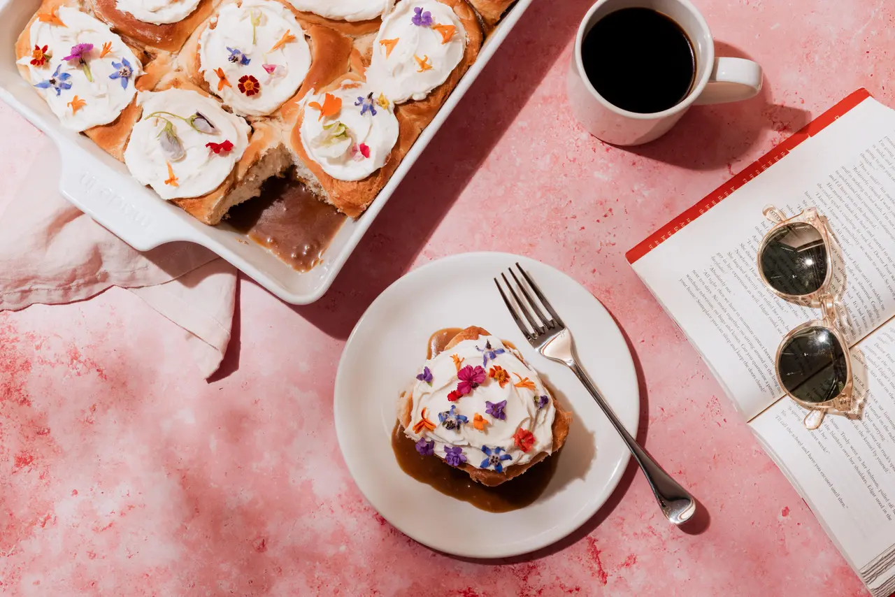 A flat lay of a breakfast setting with a plate of frosted cinnamon rolls, a cup of coffee, and reading glasses next to an open book on a pink surface.