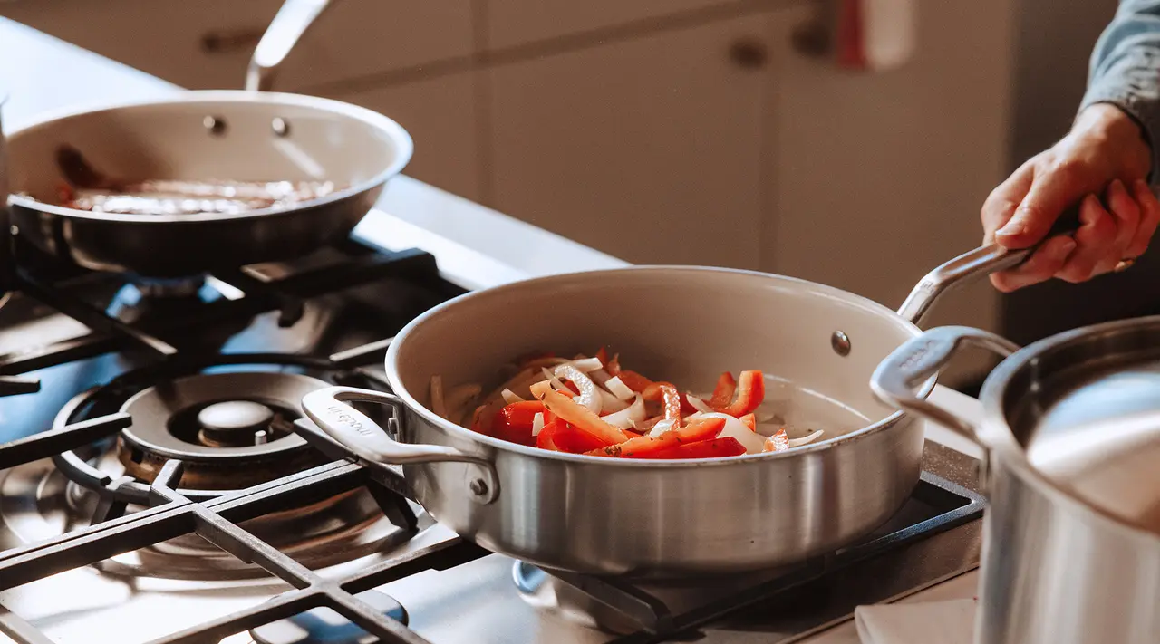 A person is cooking sliced vegetables in a silver frying pan on a stovetop, with another pan in the background.