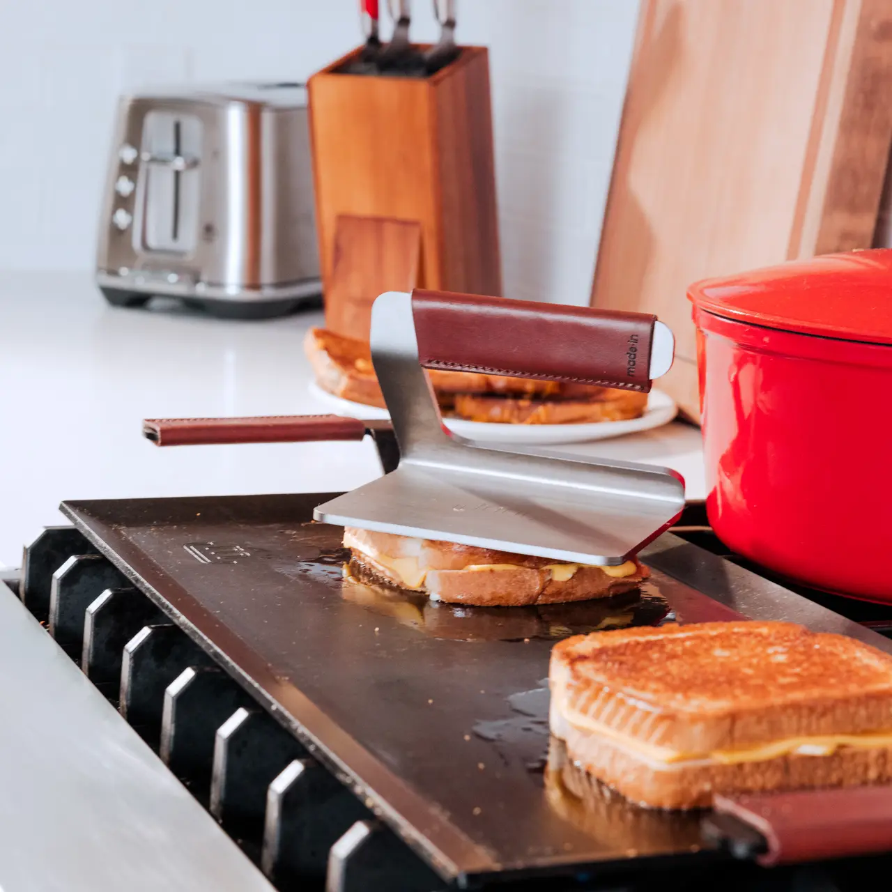 A grilled cheese sandwich is being pressed with a spatula on a stovetop griddle, with a toaster and kitchen utensils in the background.