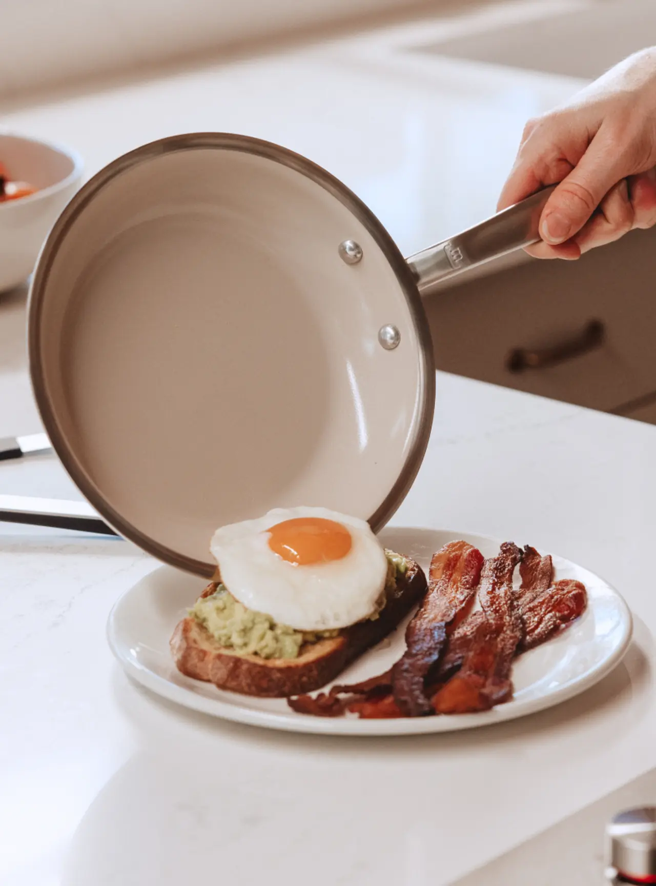 A hand is holding a frying pan above a plate with toast topped with avocado and a fried egg alongside crispy bacon.