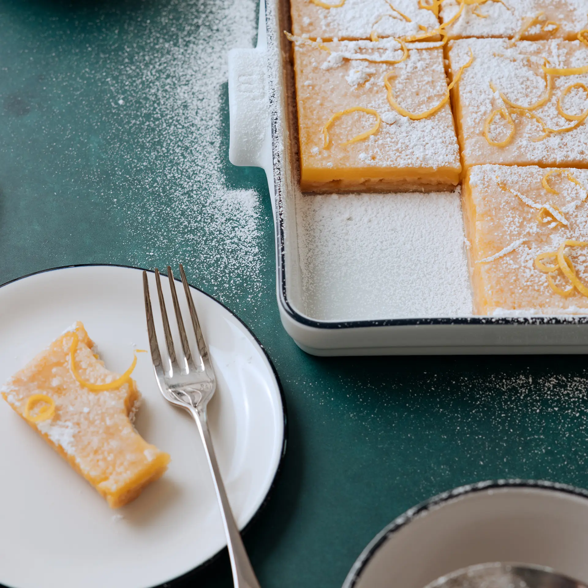 A dessert of lemon bars dusted with powdered sugar is served, with one piece on a plate and a fork nearby.