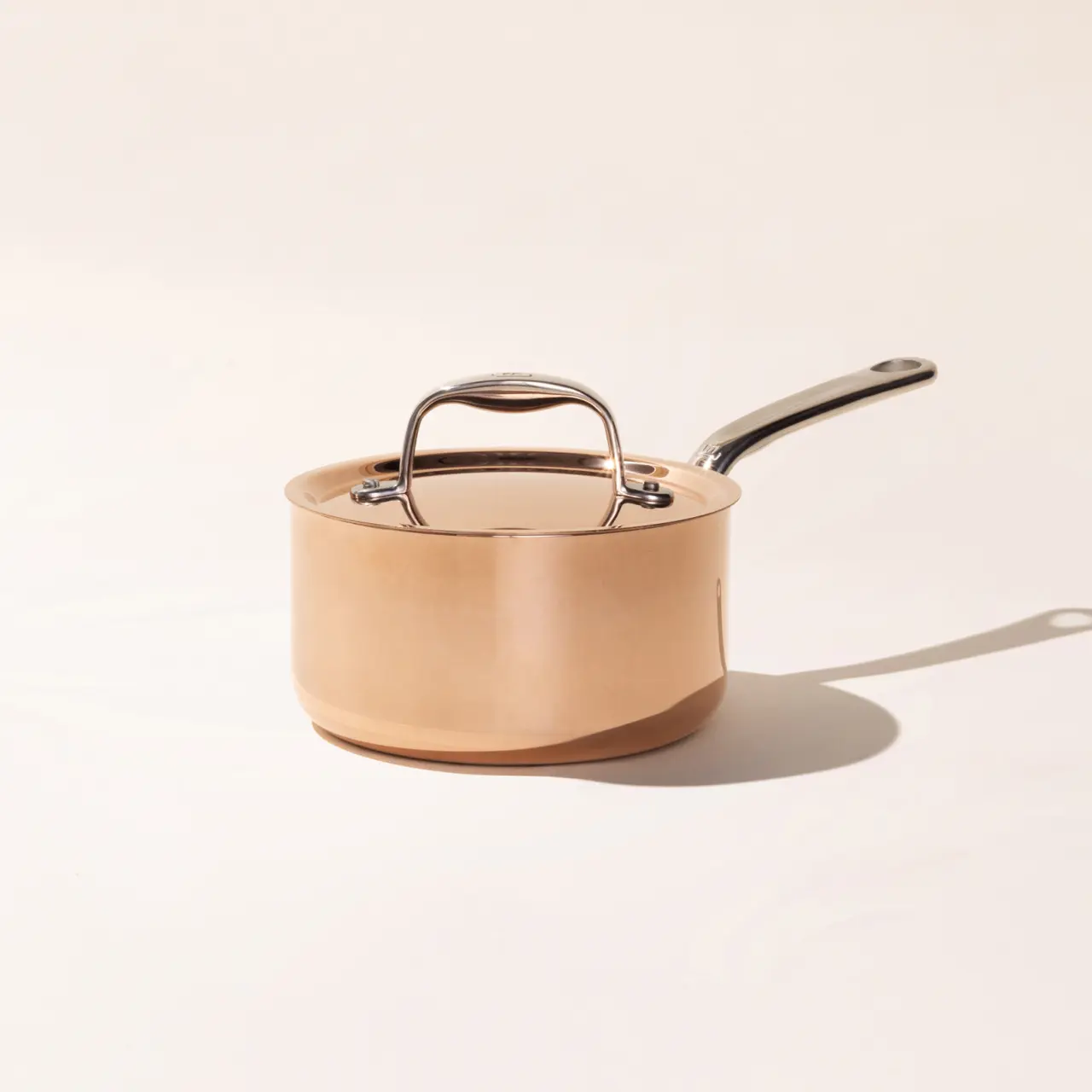 A copper saucepan with a silver handle and lid sits against a light neutral background, casting a soft shadow to the right.