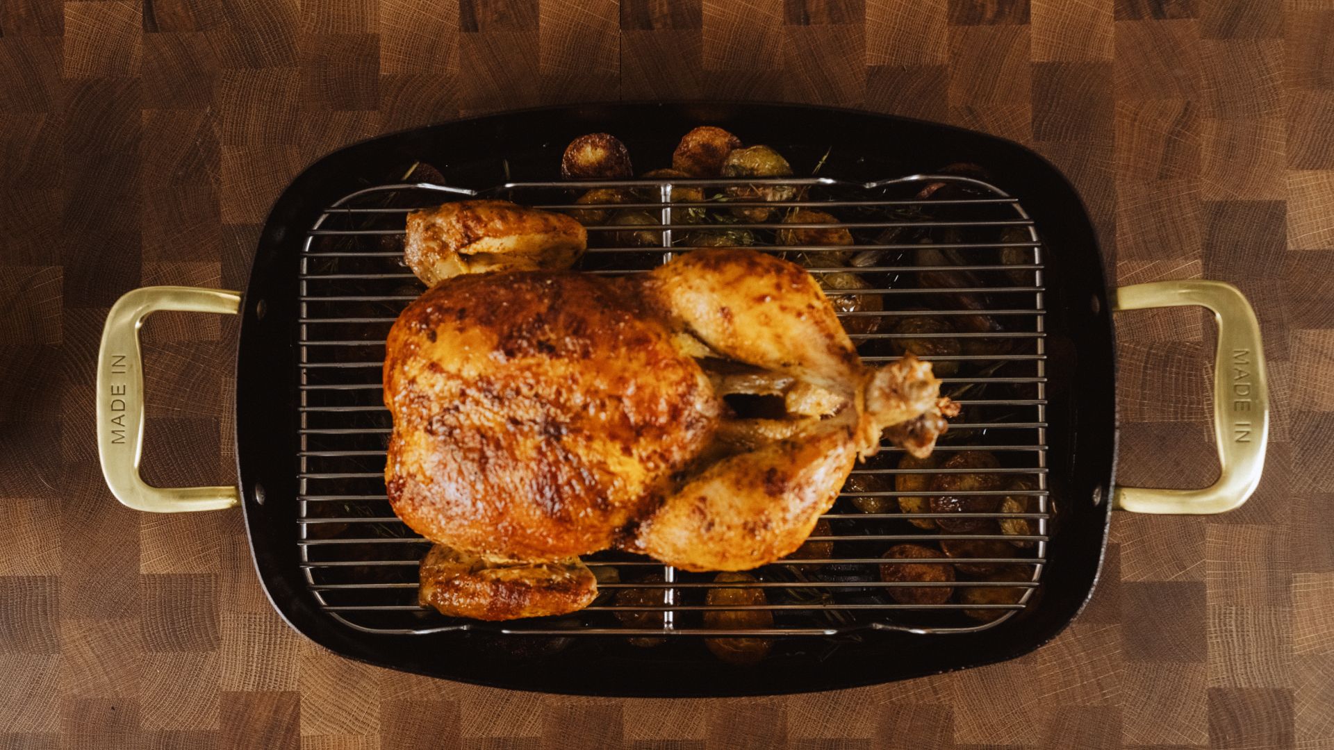 A perfectly roasted chicken sits on a rack over a tray, surrounded by small potatoes, on a wooden surface.