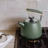 A matte green kettle with a quilted handle sits on a stove against a tiled backsplash.