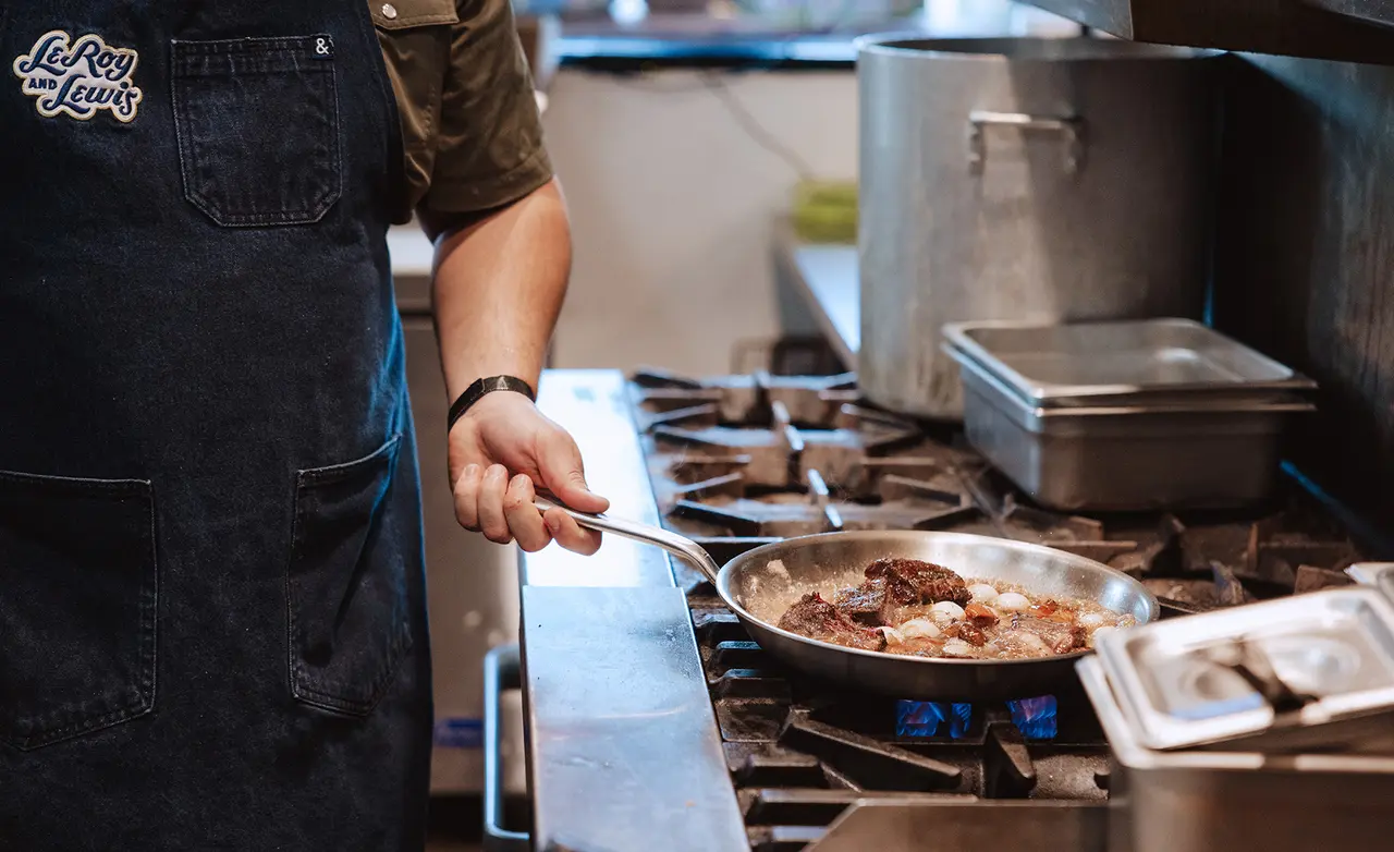 A chef in an apron is sautéing meat in a frying pan on a stove in a kitchen.