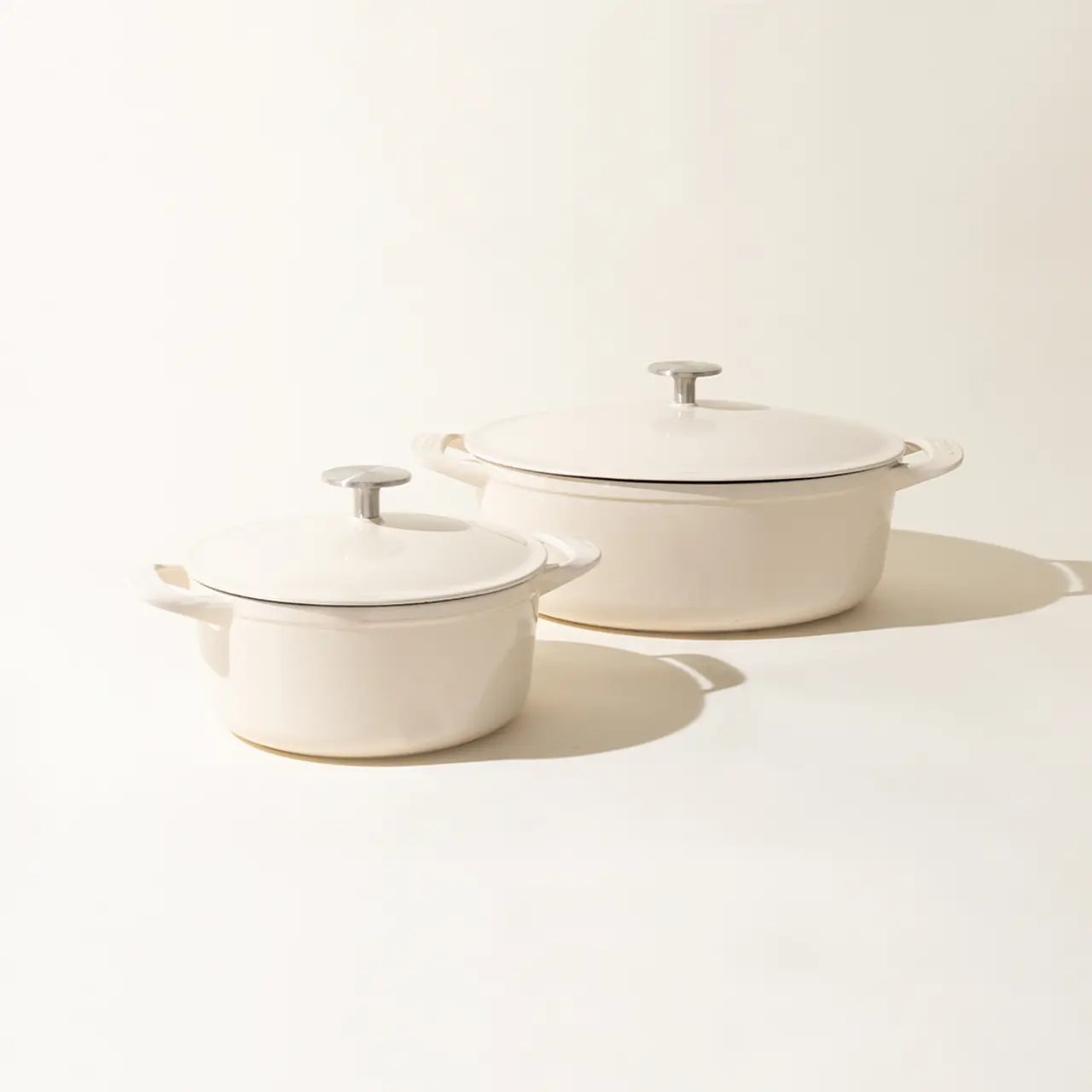 Two off-white cooking pots with lids on a light background, one larger than the other, casting soft shadows to the right.