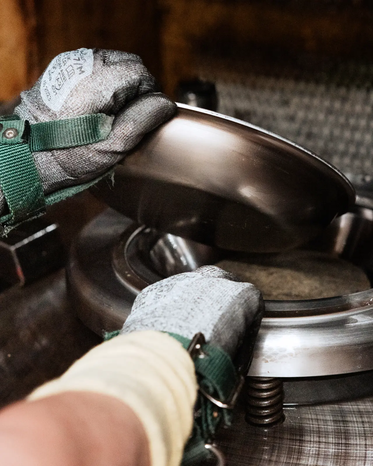 Hands wearing gloves are carefully lifting a metallic lid off a container in a workshop setting.