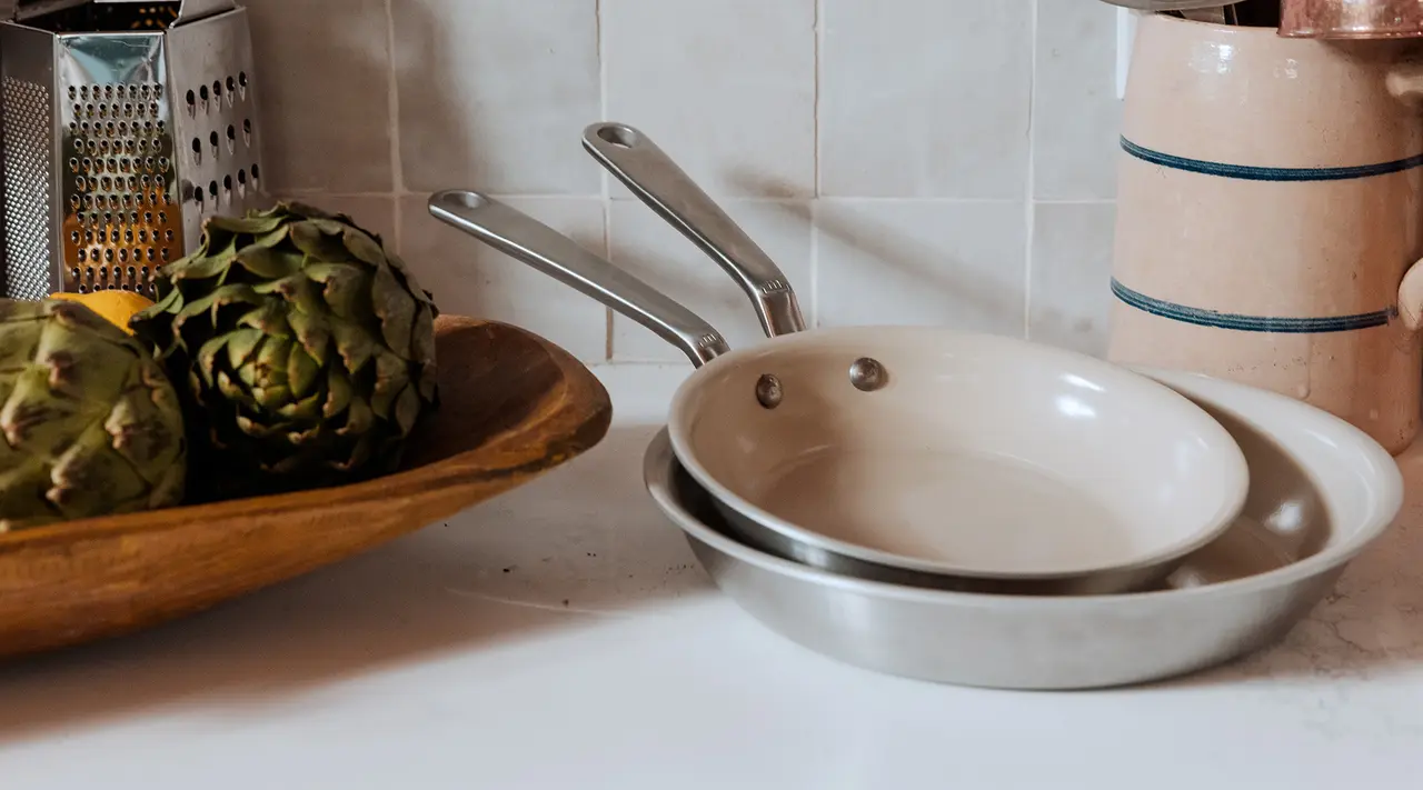 A kitchen countertop displays cooking pans and a wooden bowl containing artichokes next to a grater and a ceramic jar.