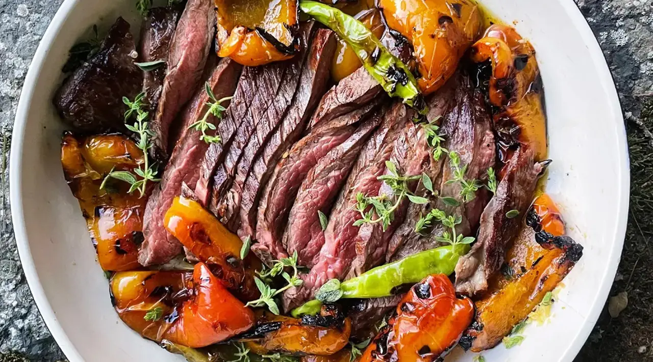 Sliced grilled steak served with roasted bell peppers and garnished with fresh herbs on a white plate.