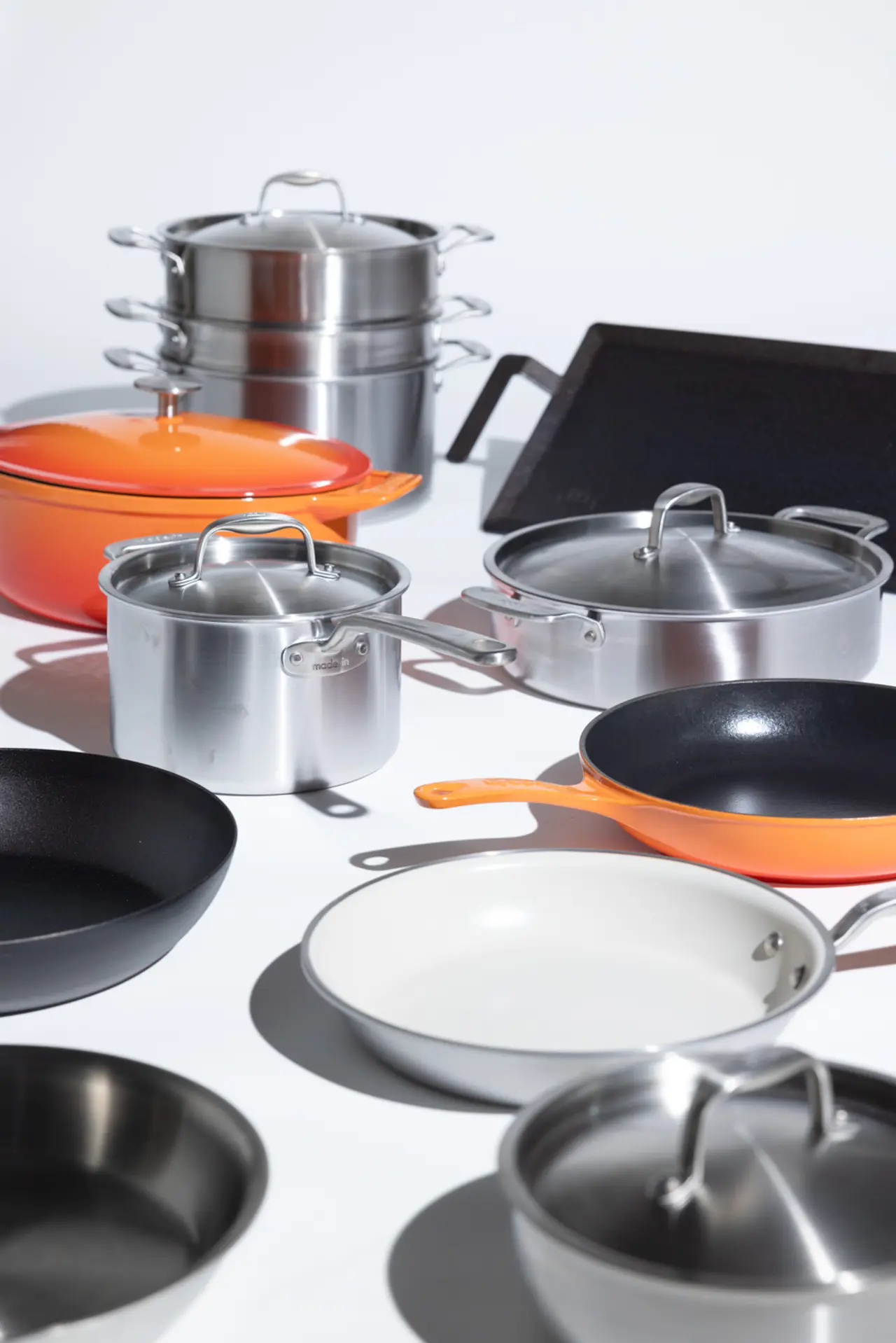 A variety of cookware, including pots and frying pans in different sizes and colors, is arranged on a white surface.