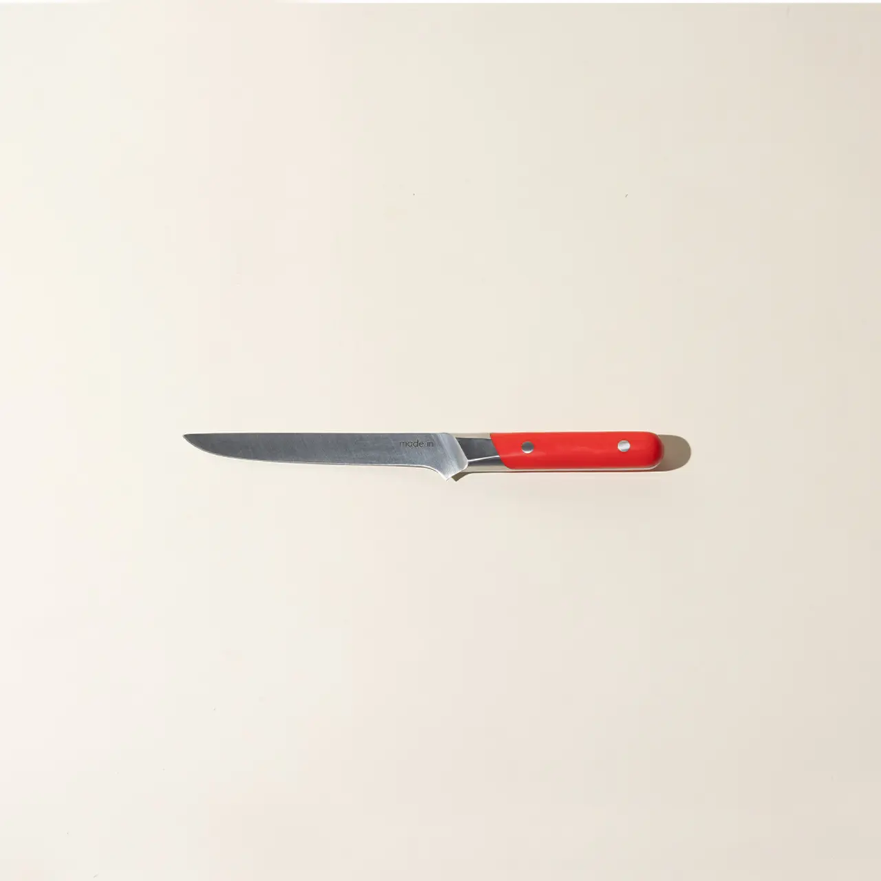 A single kitchen knife with a red handle lies centered on a plain beige background.