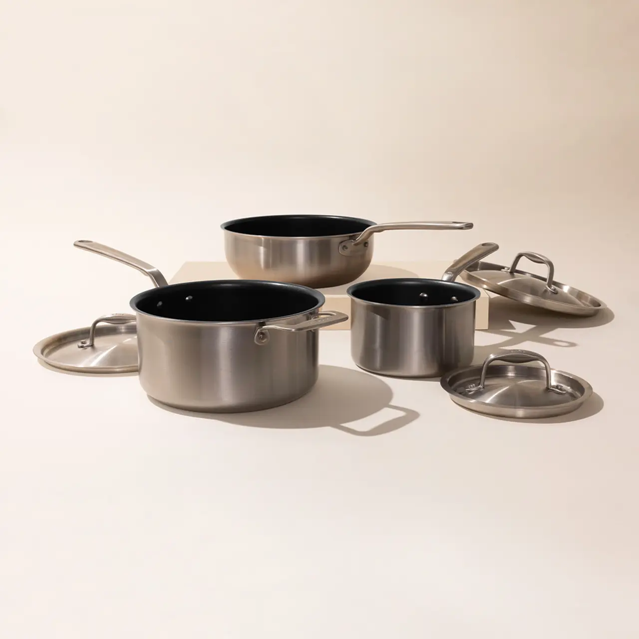 A set of three stainless steel pots with lids, featuring a large pot, a medium pot, and a small saucepan on a neutral background.