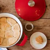 Freshly baked golden biscuits are arranged in a large white pot with a red lid, alongside a small bowl of seasoning and an individual biscuit on a white plate.