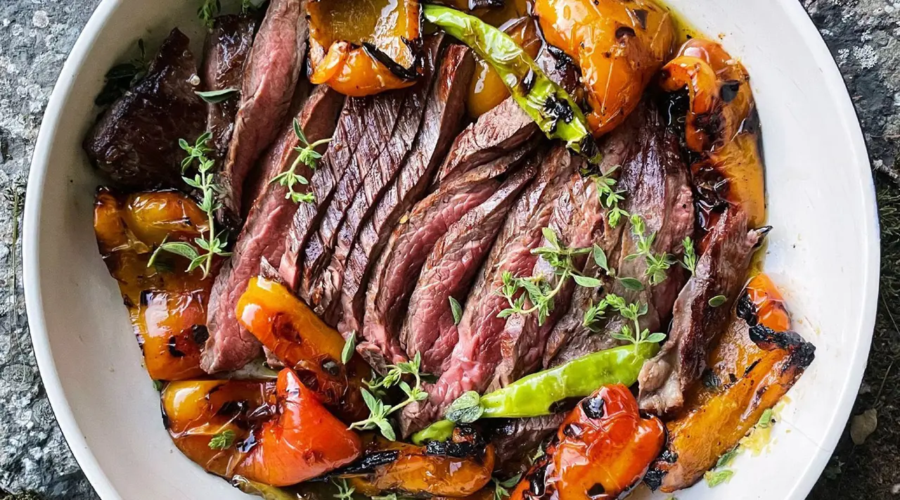 Sliced grilled steak garnished with herbs is served with charred bell peppers and asparagus on a white plate.