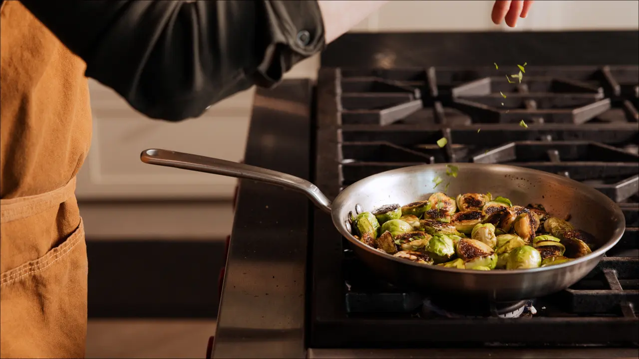 A person in an apron sautés Brussels sprouts in a stainless steel pan on a stove.