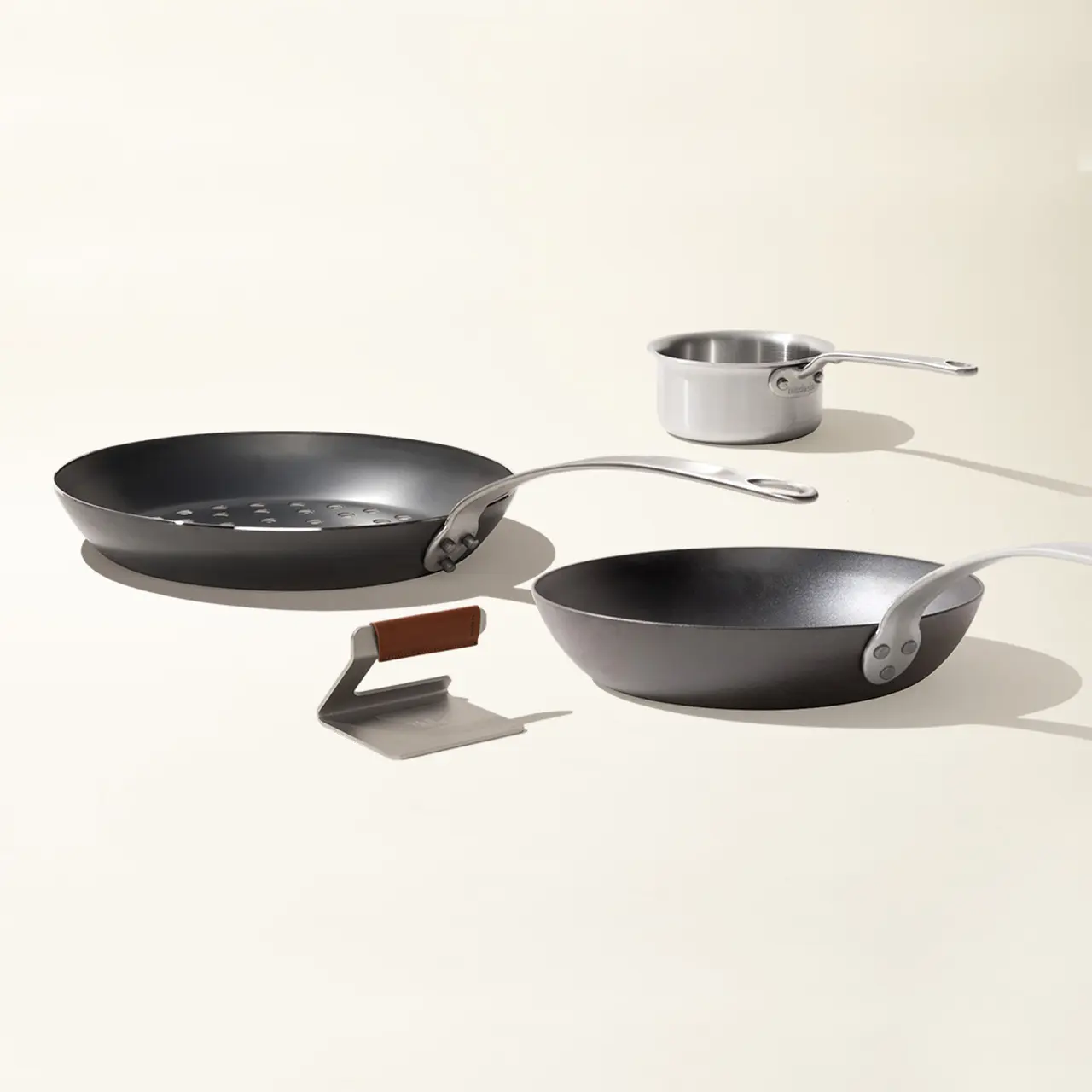 Three different-sized frying pans are arranged on a light background, accompanied by a spatula with a brown handle.