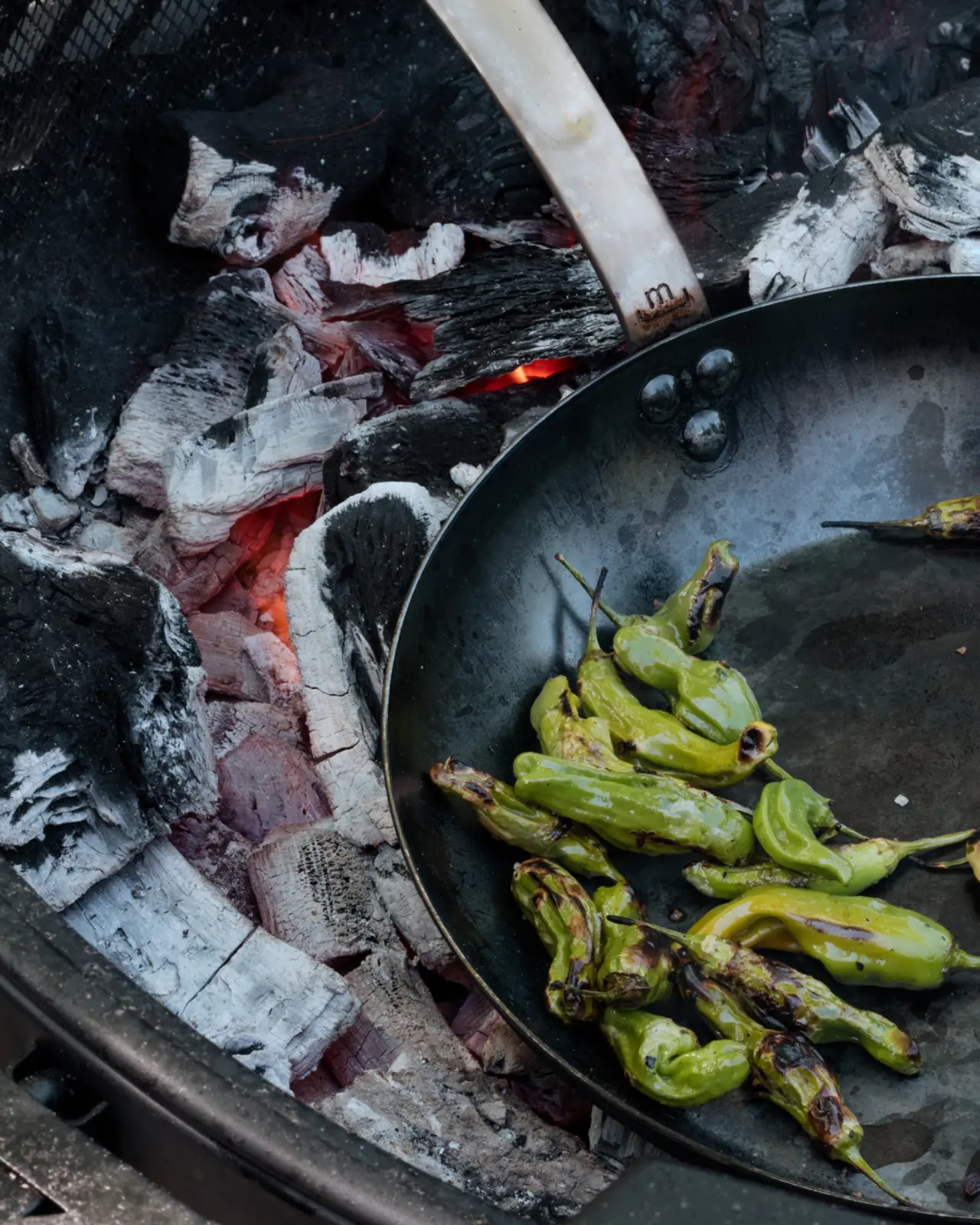 Green chili peppers are being roasted in a pan over smoldering coals.