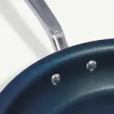 A close-up view shows the handle and part of the body of a dark-colored frying pan with rivets.