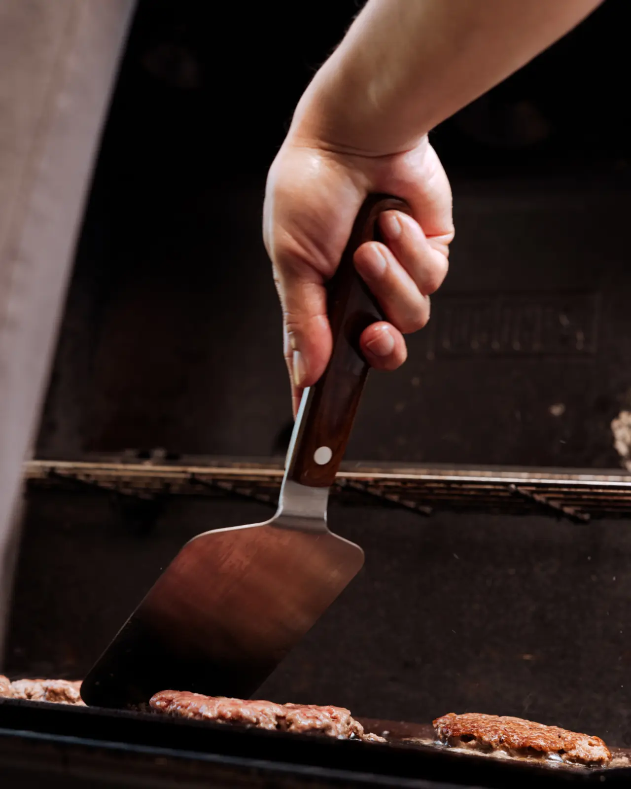 A person's hand is flipping burgers on a grill with a metal spatula.