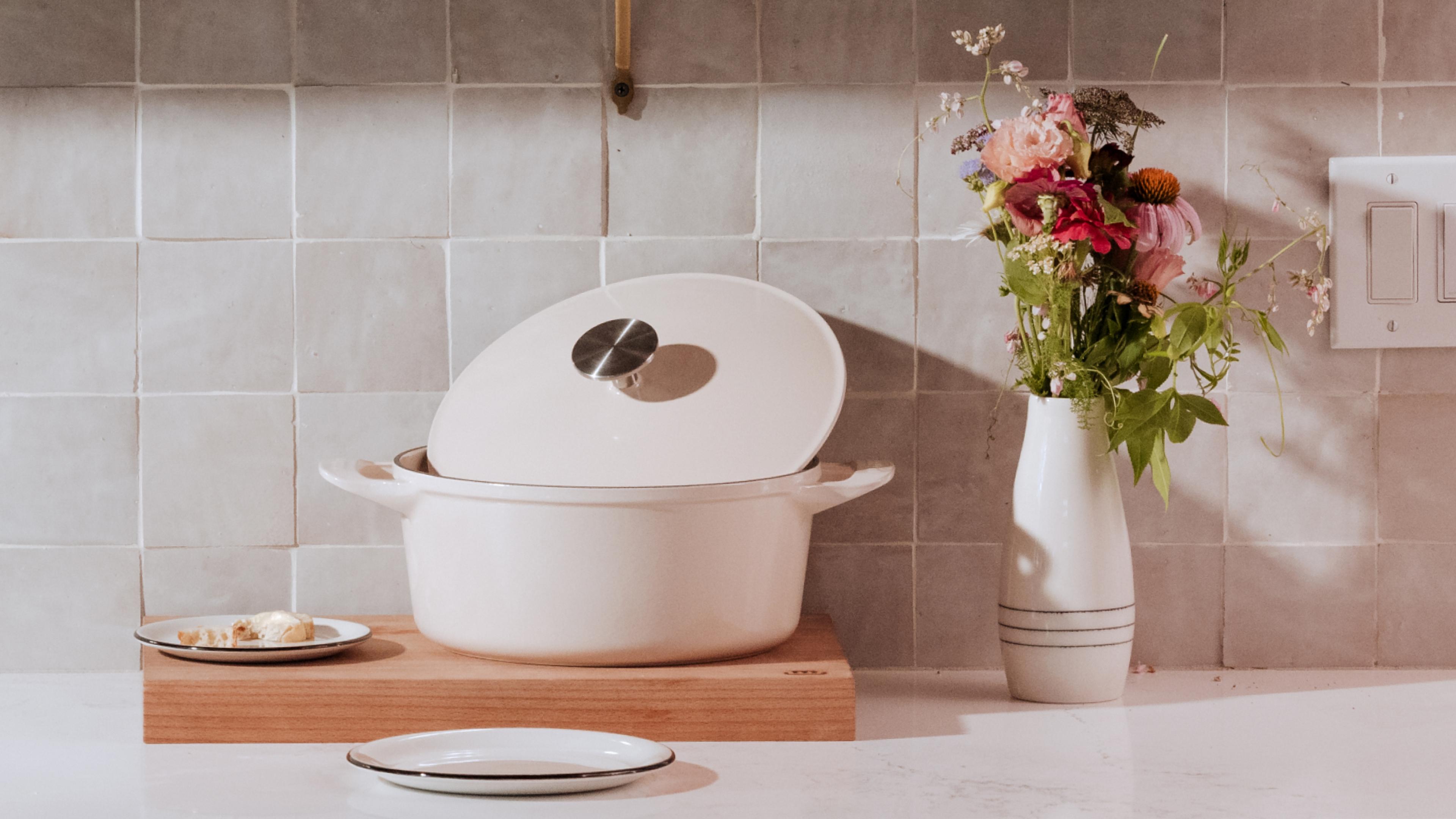 A white enameled pot rests on a wooden cutting board next to a vase of flowers on a kitchen counter.