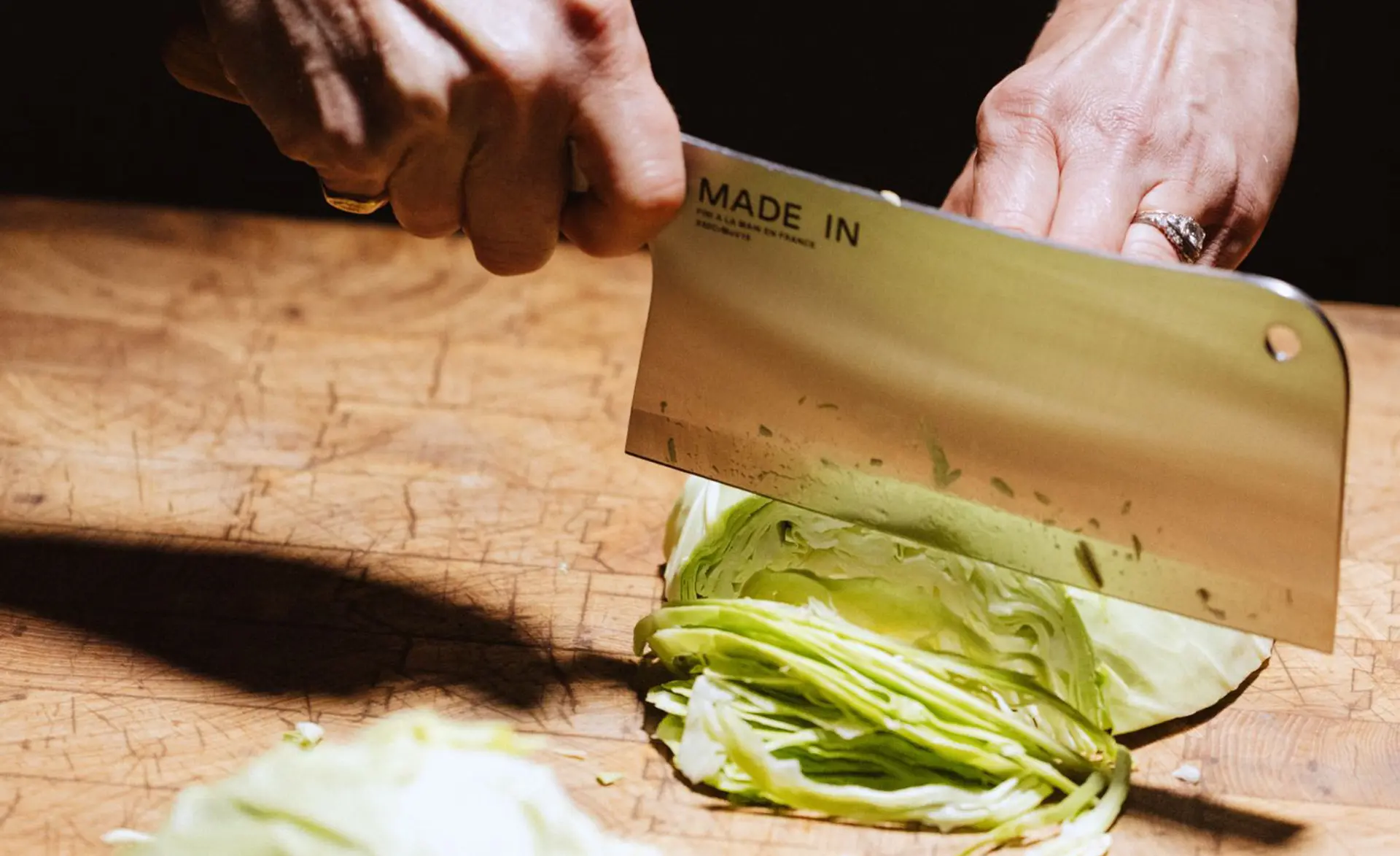A hand holds a large knife above a head of cabbage being sliced on a wooden cutting board.