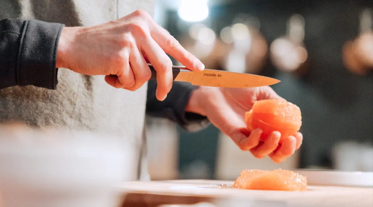 Close-up of hands using a knife to slice an orange on a cutting board.